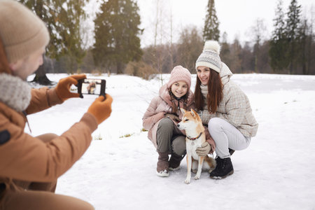 Portrait of father taking photo of cute daughter and wife with dog while enjoying walk outdoors together in winter forest, copy spaceの写真素材