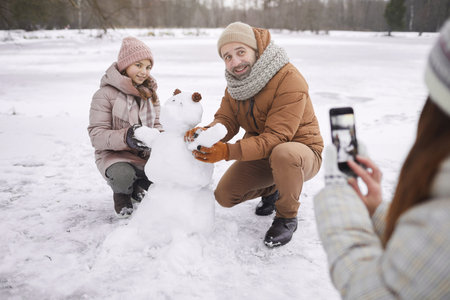 Portrait of young woman taking photo of father and daughter building snowman together while enjoying winter outdoors, copy spaceの写真素材