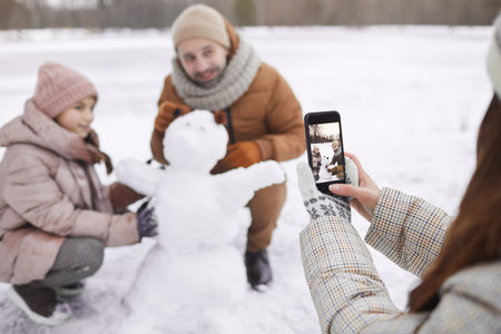 Portrait of young woman taking photo of father and daughter building snowman together, focus on smartphone screen, copy spaceの写真素材