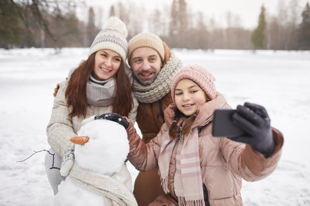 Portrait of happy family taking selfie photo outdoors while building snowman together and enjoying winter vacation, copy spaceの写真素材