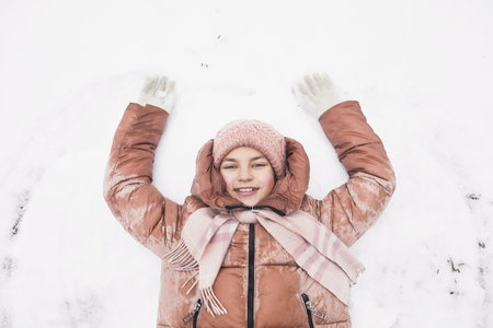 Top down view at cute girl lying in snow and making snow angels while enjoying winter outdoors and smiling at camera, copy spaceの写真素材