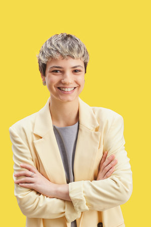 Vertical waist up portrait of confident short-haired woman smiling at camera while standing with arms crossed against pop yellow backgroundの写真素材