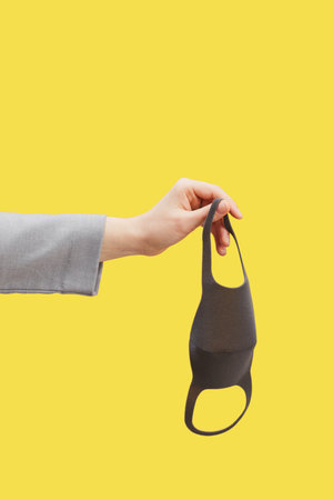 Graphic image of female hand holding mask against pop yellow background, copy space