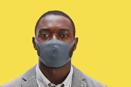 Head and shoulders portrait of African-American man wearing mask and looking at camera while standing against pop yellow background, copy spaceの写真素材