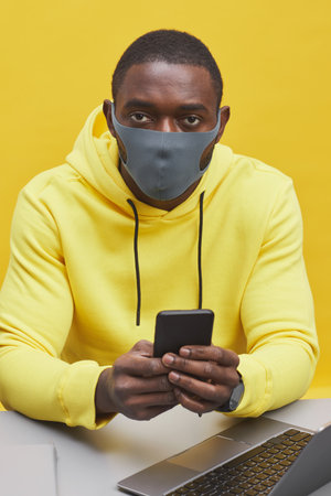 Vertical portrait of contemporary African-American man wearing mask while sitting at desk against pop yellow backgroundの写真素材
