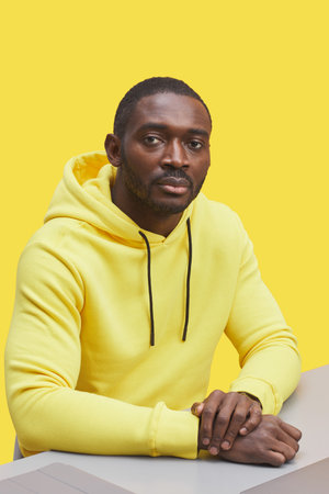 Vertical portrait of contemporary African-American man wearing hoodie and looking at camera while sitting at desk against illuminating yellow backgroundの写真素材