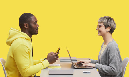 Side view portrait of two contemporary business people sitting opposite each other at desk during meting and smiling against yellow backgroundの写真素材