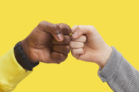 Graphic close up of two young people bumping fists against illuminating yellow background, interracial friendship and unity concept, copy spaceの写真素材