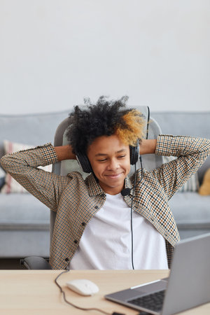Vertical portrait of smiling African-American boy wearing headset relaxing in chair while using laptop at homeの写真素材