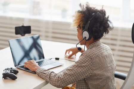 Side view portrait of teenage African-American boy playing video games at home and online streaming,...