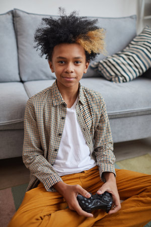 Vertical portrait of smiling African-American boy playing video games at home and looking at camera...
