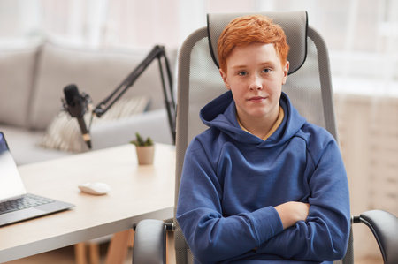 Portrait of red haired teenage boy looking at camera while sitting in chair against computer in background, copy spaceの写真素材