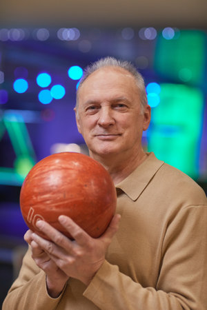 Vertical waist up portrait of smiling senior man holding bowling ball and looking at camera while enjoying active entertainment at bowling alleyの写真素材