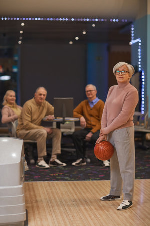 Vertical full length portrait of group of senior people playing bowling with focus on mature woman holding bowling ball in foreground, copy spaceの写真素材