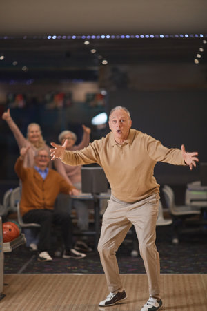 Vertical full length portrait of excited senior man playing bowling and celebrating win or strike with group of friends in backgroundの写真素材