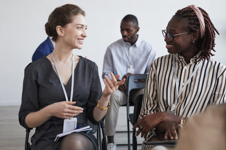 Portrait of two smiling businesswomen talking while sitting on chairs in audience at business...
