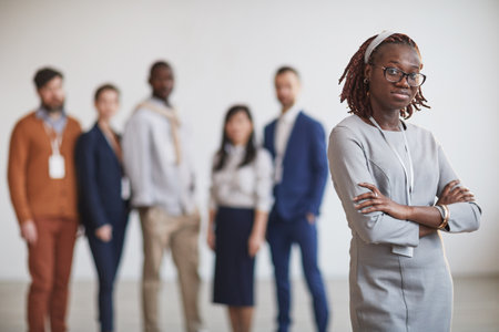 Waist up portrait of successful African-American businesswoman standing with arms crossed against multi-ethnic team in background, copy spaceの写真素材