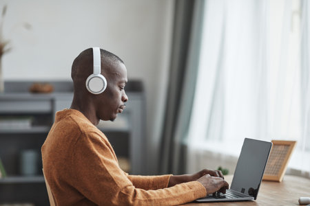 Side view portrait of African-American man using laptop at desk while working from home in minimal...