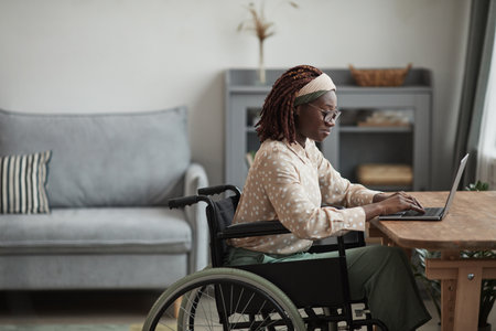 Side view portrait of young African-American woman using wheelchair while working from home in minimal grey interior, copy spaceの写真素材