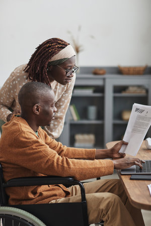 Vertical side view portrait of African-American man using wheelchair working from home with wife looking over his shoulder, copy spaceの写真素材