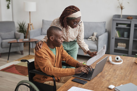 Portrait of African-American man using wheelchair working from home with wife looking over his...