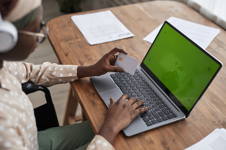 High angle close up of African-American woman in wheelchair online shopping via laptop and holding credit card to green screen, copy spacの写真素材