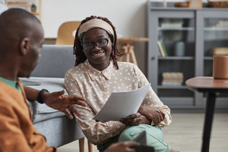 Portrait of modern African-American couple working from home together, focus on smiling young woman holding document while sitting on floor, copy spaceの写真素材