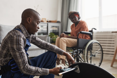 Side view portrait of African-American man assembling furniture in home interior with woman in wheelchair in background, handyman service and assistance conceptの写真素材