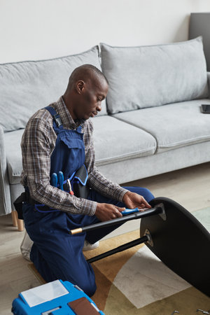 Vertical side view portrait of African-American handyman assembling furniture in home interior, service and assistance conceptの写真素材