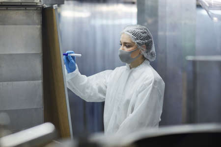 Portrait of female worker wearing mask and protective suit while writing on board in factory...