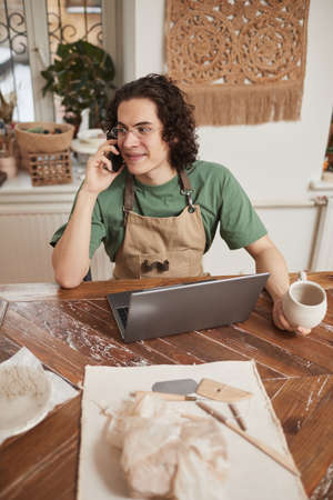 Vertical portrait of young man speaking by smartphone and using laptop in workshop while managing small business, copy spaceの写真素材