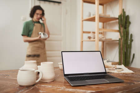 Still life of laptop with blank white screen on table in workshop, with blurred male artisan speaking by phone in background, small business and entrepreneurship concept, copy spaceの写真素材