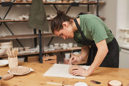 Side view portrait of young man decorating ceramic bowl in pottery workshop, hobby and small...