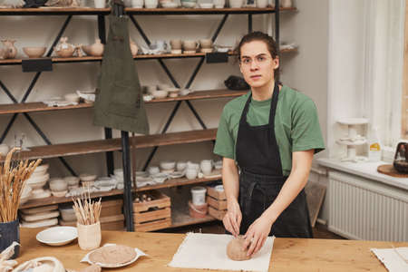Warm toned portrait of young man looking at camera while making ceramic bowl in pottery workshop,...