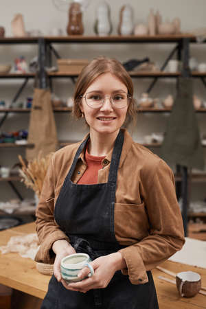 Vertical waist up portrait of young female potter looking at camera while posing in workshop, hobby...