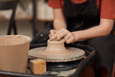 Warm-toned close up of female hands shaping clay on pottery wheel in workshop while enjoying arts and crafts, copy spaceの写真素材