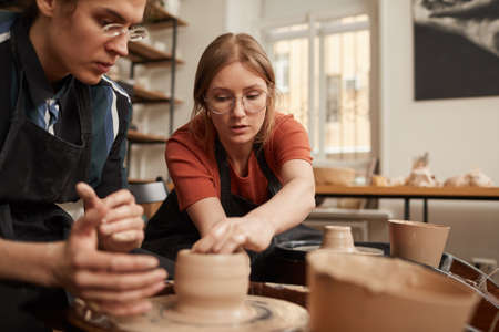 Portrait of female potter helping young man shaping clay on pottery wheel in workshop, copy spaceの写真素材