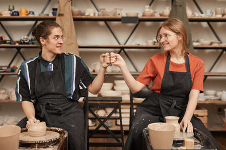 Front view portrait of two young artisans bumping fists while making ceramics in pottery workshop with pottery wheel, copy spaceの写真素材