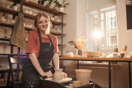 Warm toned portrait of young female artisan smiling at camera while working on pottery wheel in sunlit workshop and enjoying arts and crafts, copy spaceの写真素材
