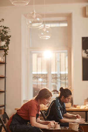 Vertical side view portrait of two young people shaping clay on pottery wheel while making ceramics...