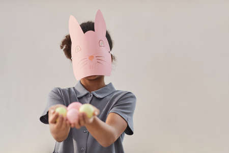 Minimal waist up portrait of African-American teenager wearing bunny mask and holding Easter eggs...
