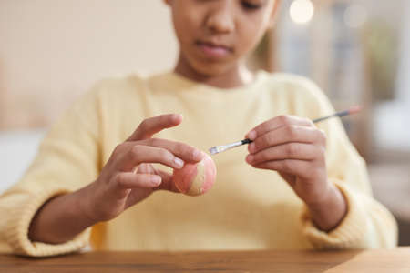 Close up of young African-American girl hand painting Easter eggs with pastel colors while enjoying...
