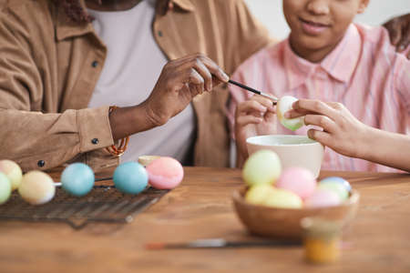 Close up of loving African-American mother and daughter painting Easter eggs together while sitting at wooden table in cozy home interiorの写真素材
