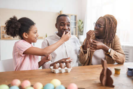 Portrait of loving African-American family making chocolate Easter decorations and tasting them while sitting at wooden table in cozy home interiorの写真素材