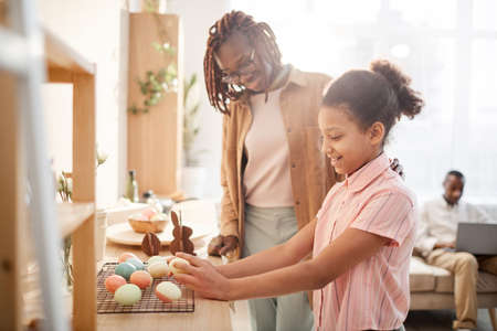 Side view portrait of smiling African-American girl preparing Easter decorations with mom in cozy home interior, copy spaceの写真素材