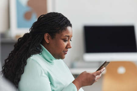 Side view portrait of curvy African American woman looking on smartphone screen while browsing...