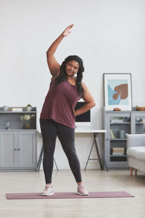 Vertical full length portrait of curvy African American woman working out at home and smiling at...