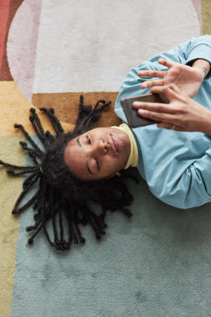 Top view portrait of real African American woman using smartphone or taking selfie while lying on floor on colorful rug, , focus on skin imperfectionsの写真素材