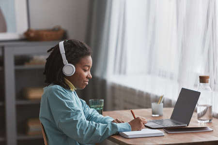 Side view portrait of young African American woman studying or working at home and smiling while wearing headphones, copy spaceの写真素材