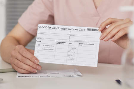 Close up of young female nurse holding vaccination record card while registering patients for covid...